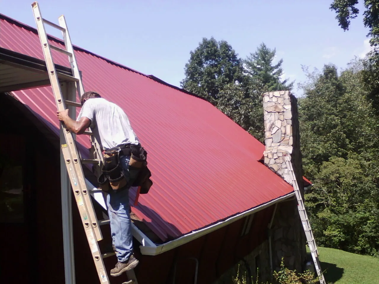 Roofer on a ladder beside a red metal roof and stone chimney — Soffit & Fascia Repair by Roofing Brothers in Nome
